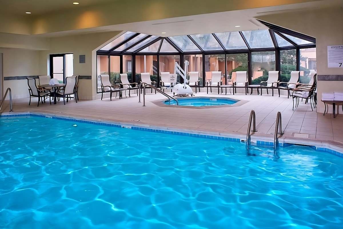 An indoor swimming pool area features clear blue water, surrounded by spacious seating options including loungers and dining tables. Large windows provide natural light and views of the outdoors, enhancing the inviting atmosphere of the space.
