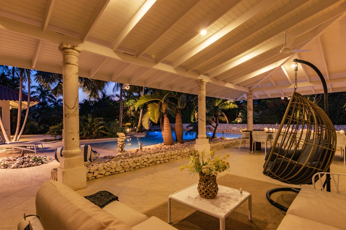 An outdoor living area is presented with a hanging chair and comfortable seating. The space is complemented by stone columns and a view of lush palm trees beside a softly lit pool. Ambient lighting enhances the serene atmosphere of the porch.