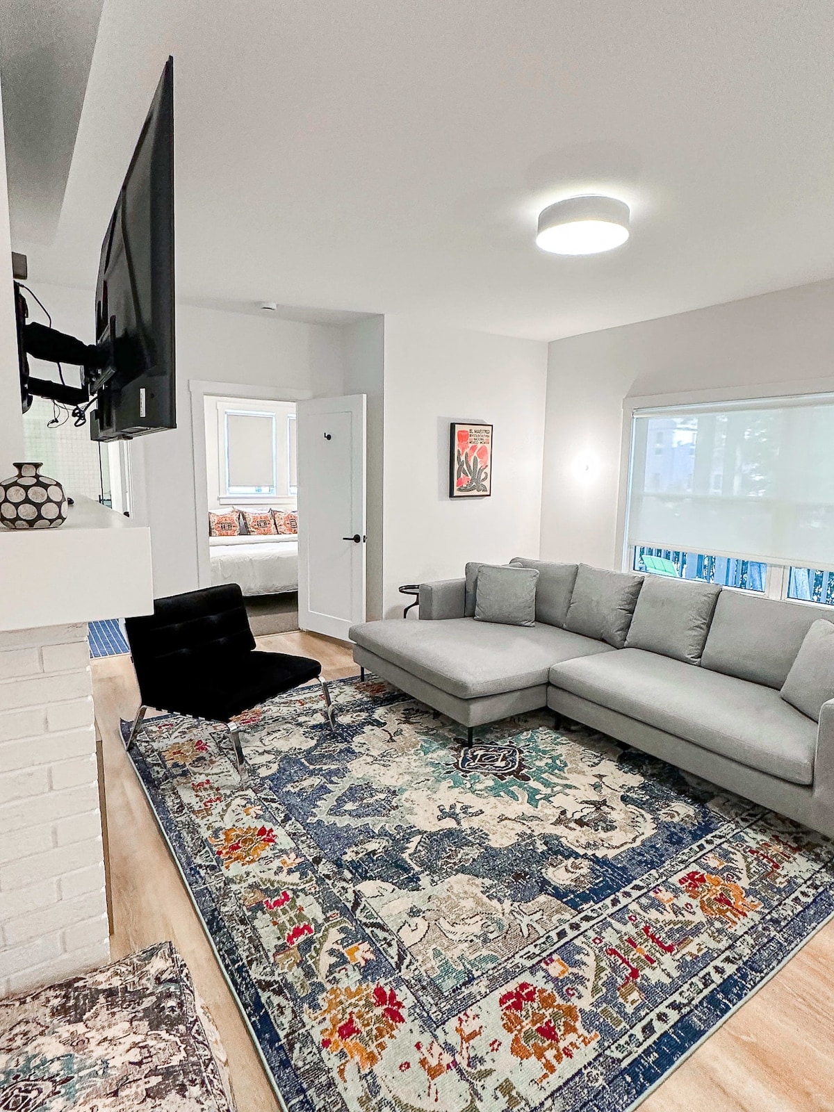 A modern living area features a light gray sectional sofa and a stylish black chair, placed atop a colorful area rug. A flat-screen TV is mounted on the wall, and natural light enters through large windows, creating a bright atmosphere.