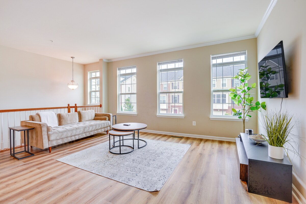 A spacious living room is designed with natural light flowing in through multiple large windows. A comfortable sofa sits alongside a round coffee table on a light area rug, and a modern television is mounted on the wall. Decorative plants add a fresh touch to the space.