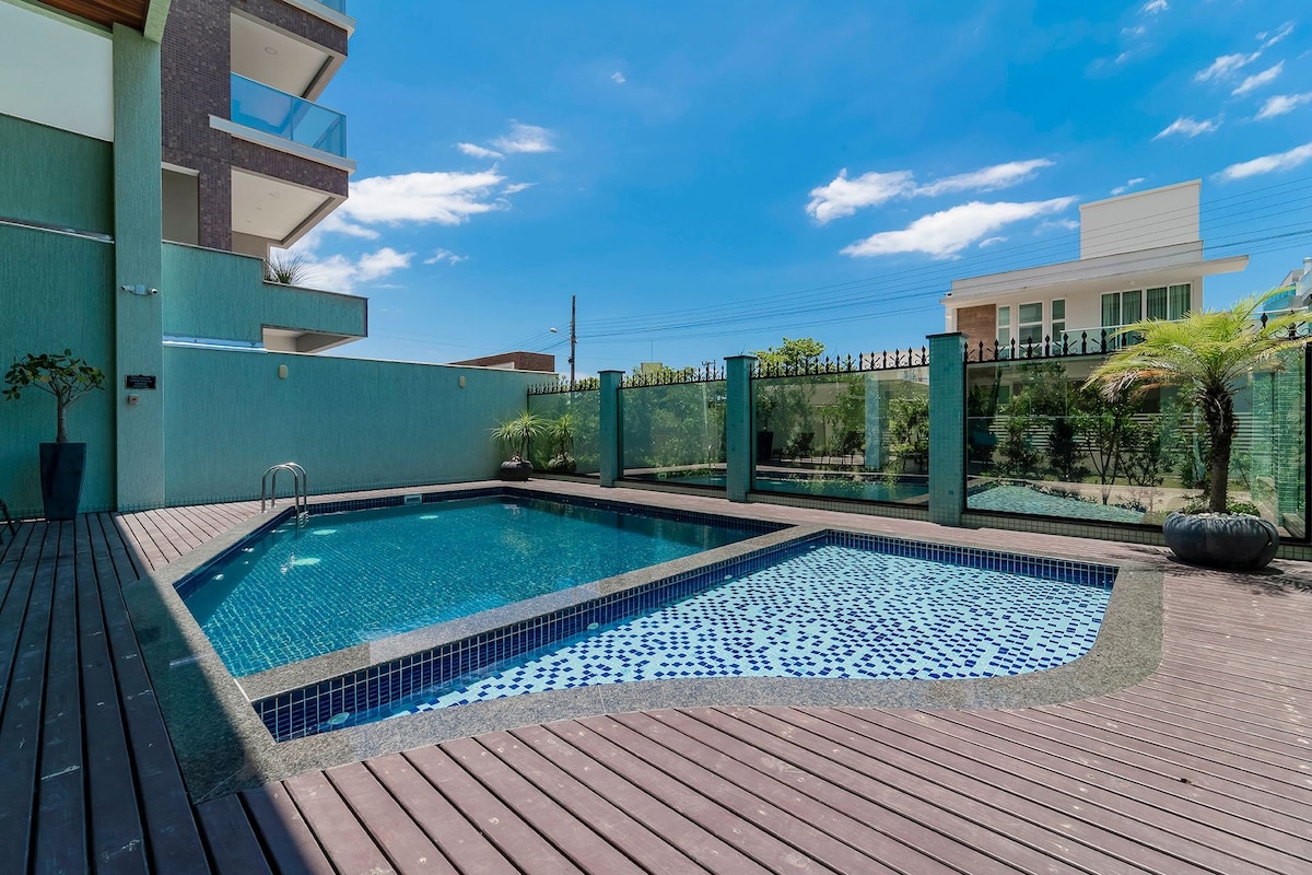 A swimming pool is presented, surrounded by wooden decking and enclosed by glass fencing. Plants are visible along the perimeter, contributing to a serene environment. The clear blue water reflects the bright sky, creating a refreshing and inviting outdoor space.