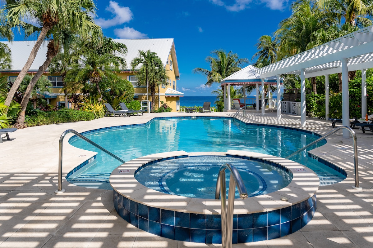 A serene outdoor area features a sparkling swimming pool and a nearby hot tub, surrounded by lush tropical landscaping. Sun loungers are positioned around the pool, and a shaded pergola provides relaxation space. The ocean is visible beyond the pool area under a clear blue sky.