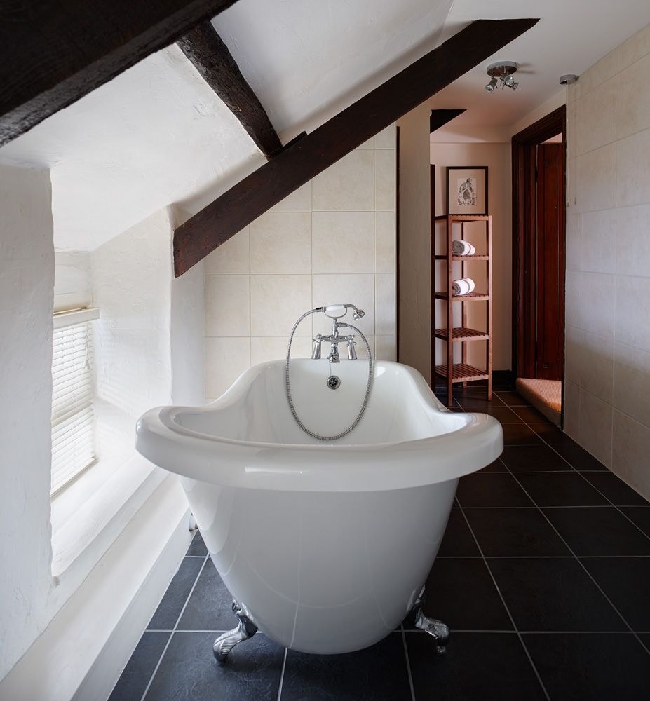 A stunning slipper bathtub is positioned under an angled ceiling, featuring ornate chrome fixtures. Natural light filters through a nearby window, illuminating the light-colored tiled walls and dark floor. Wooden beams add a touch of character to the serene bathroom space.