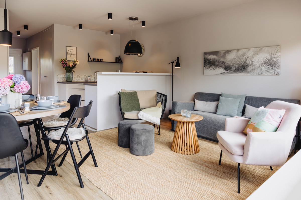 A spacious living and dining area features a round table set with dishes and flowers. Comfortable seating includes a gray sofa, a pink armchair, and a round coffee table. Soft textures, including a rug and cushions, enhance the inviting environment.