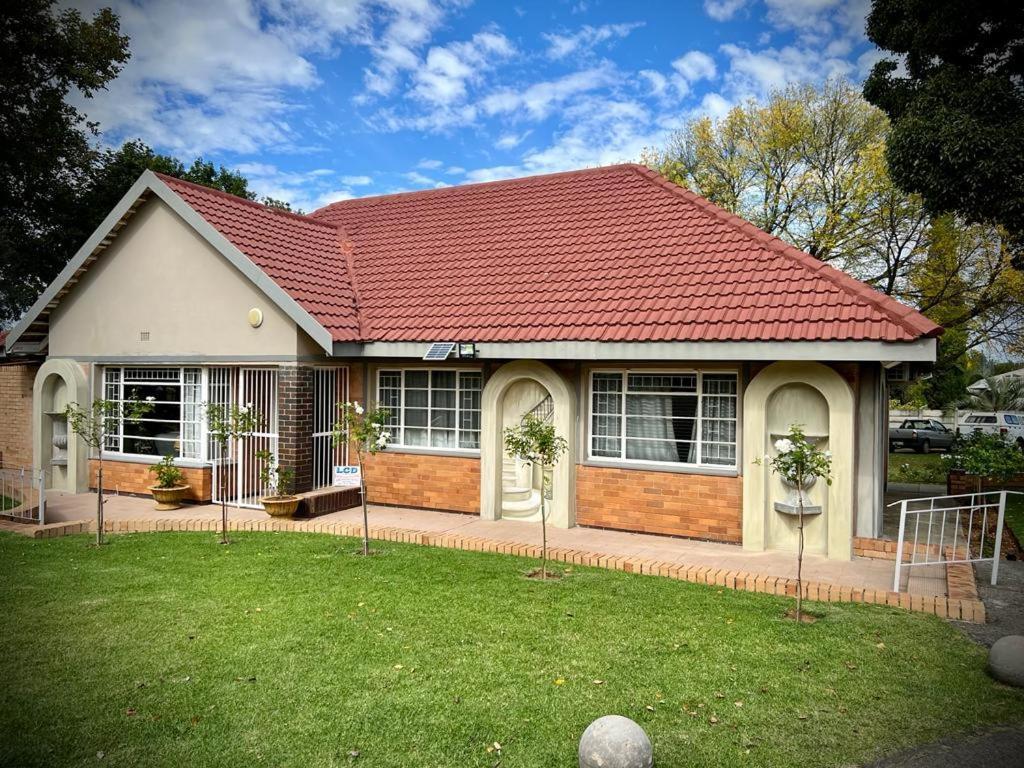 A single-story house is presented, featuring a terracotta-tiled roof and multiple rounded doorways. The front yard is green and well-maintained, with small trees lining the pathway. Large windows allow natural light to enter, creating a welcoming exterior.