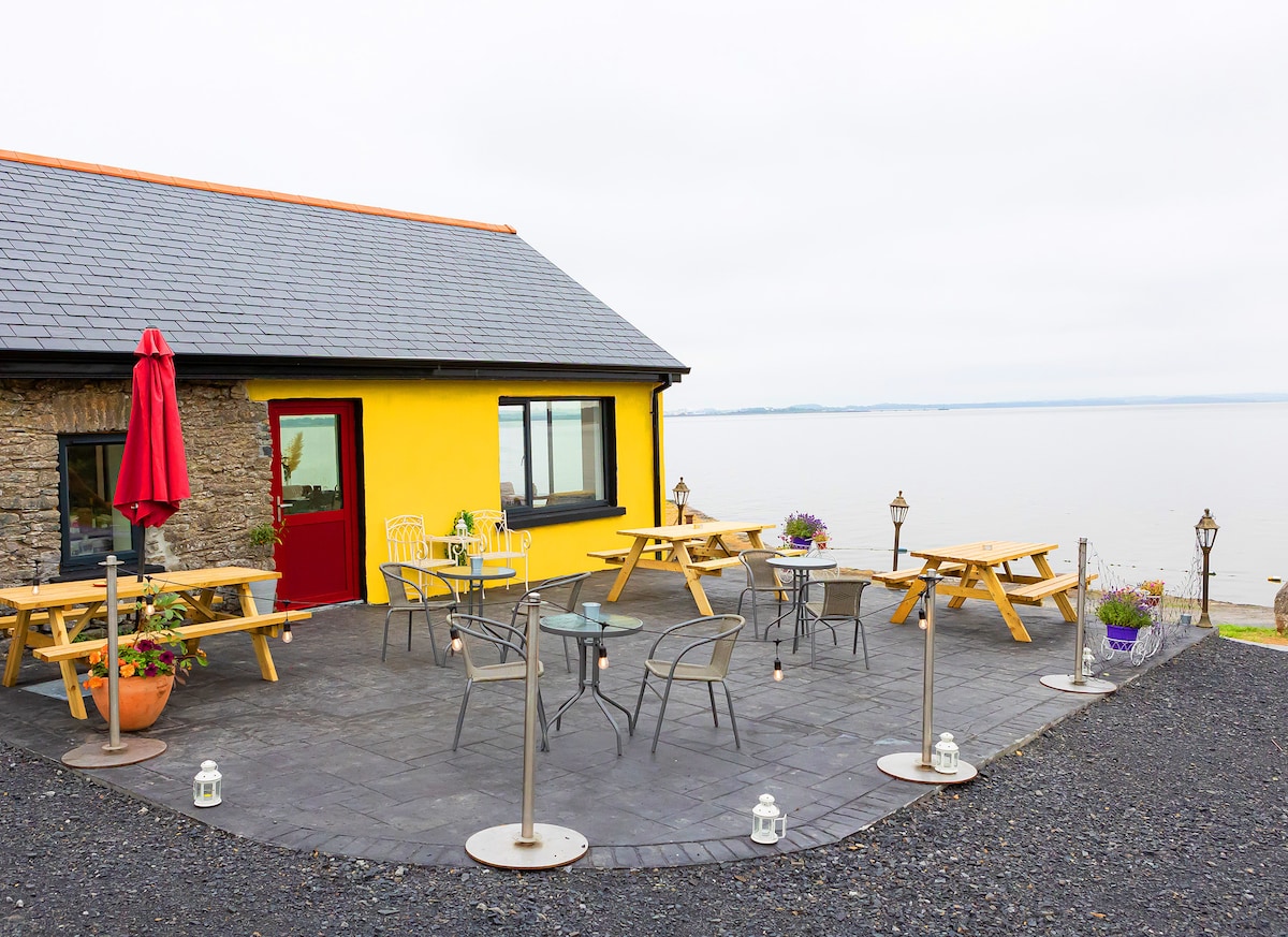 A spacious outdoor terrace is shown, featuring multiple picnic tables and chairs. A vibrant yellow wall contrasts with the stone exterior. Lanterns are arranged on the ground, and flower pots add touches of greenery. The serene waters of the Shannon Estuary are visible in the background.
