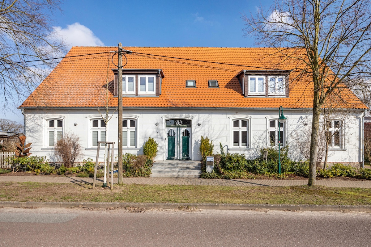 The exterior of the holiday apartment features a white structure with a red-orange pitched roof. Two dormer windows are visible at the top. The entrance is marked by two green doors, framed by greenery and neatly arranged shrubs. A pathway leads up to the entrance.