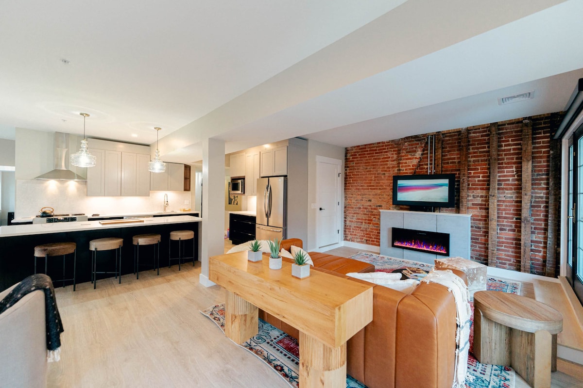An open living space is showcased with a combination of modern and vintage elements. A wooden dining table sits in the center, surrounded by comfortable seating. Exposed brick walls add character, while a sleek fireplace and a mounted television provide focal points within the area.