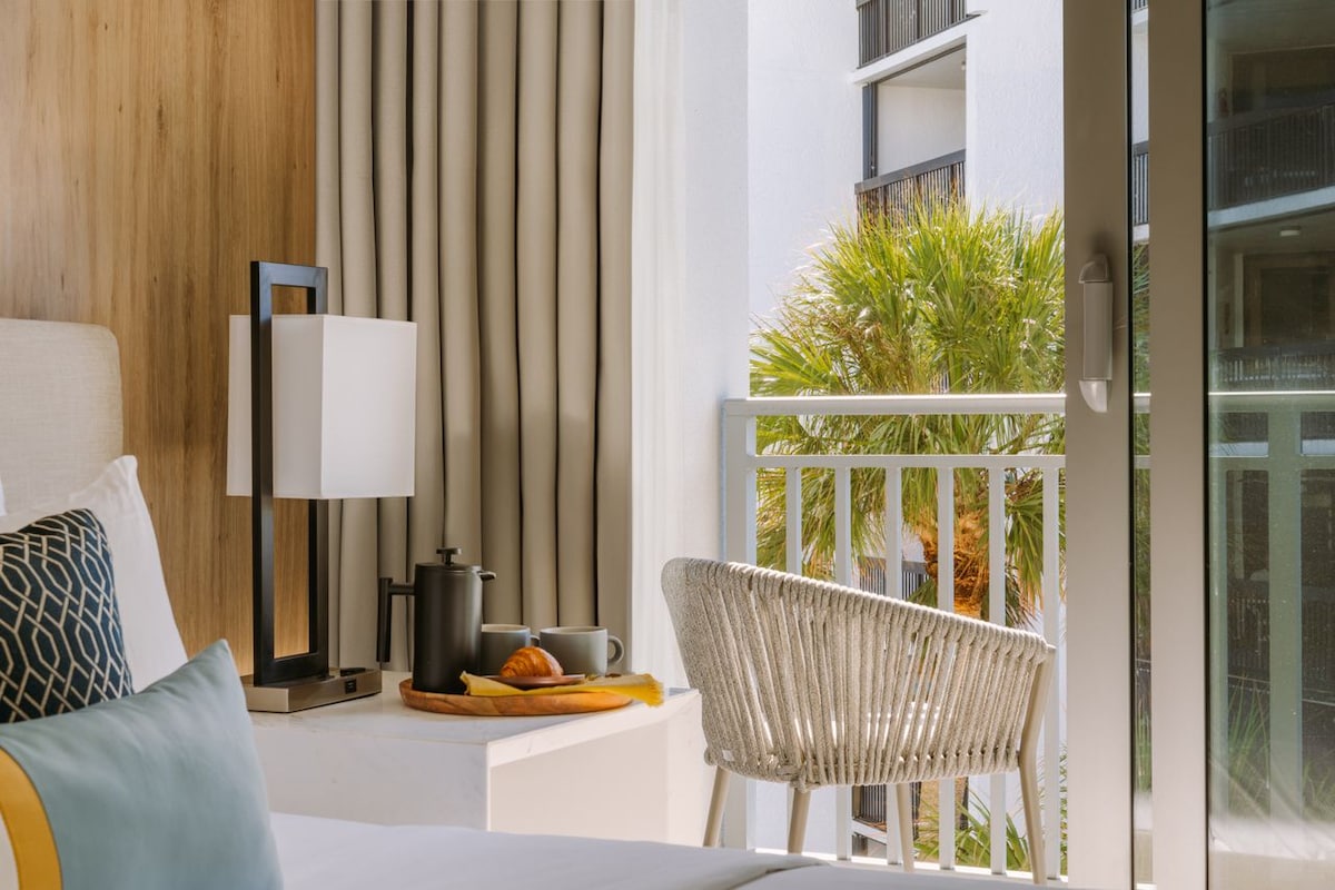 A bedside table with a modern lamp and a tray holds a teapot and croissant, providing a cozy breakfast setting. Natural light fills the room through a sliding glass door, which opens to a private balcony featuring a palm tree.