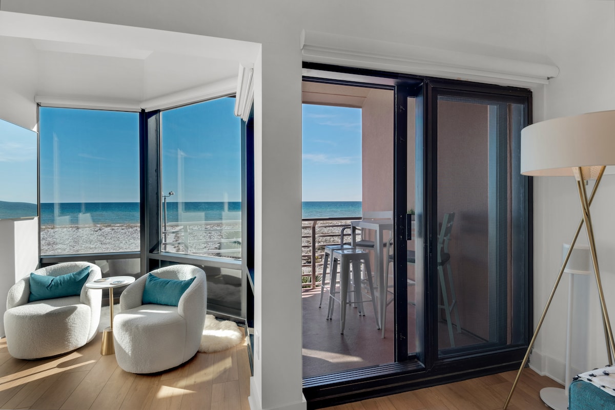 A bright, open space features large glass doors leading to a balcony with views of the Gulf of Mexico. Two cozy chairs with turquoise cushions are positioned beside a white round table, enhancing the serene coastal ambiance.