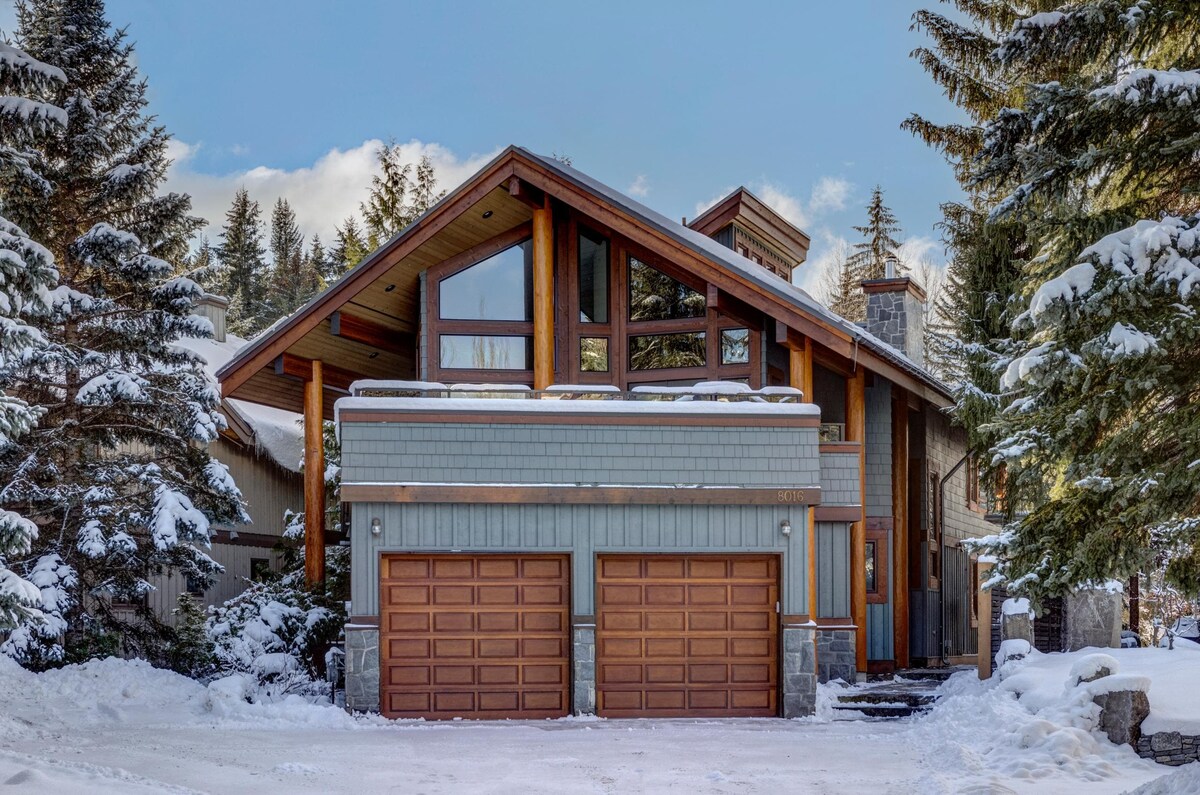 The exterior of the chalet features a modern architectural design with a steeply pitched roof and large windows. The façade includes natural wood and stone elements, while two garage doors provide convenient access. Snow blankets the ground and trees, creating a serene winter landscape.