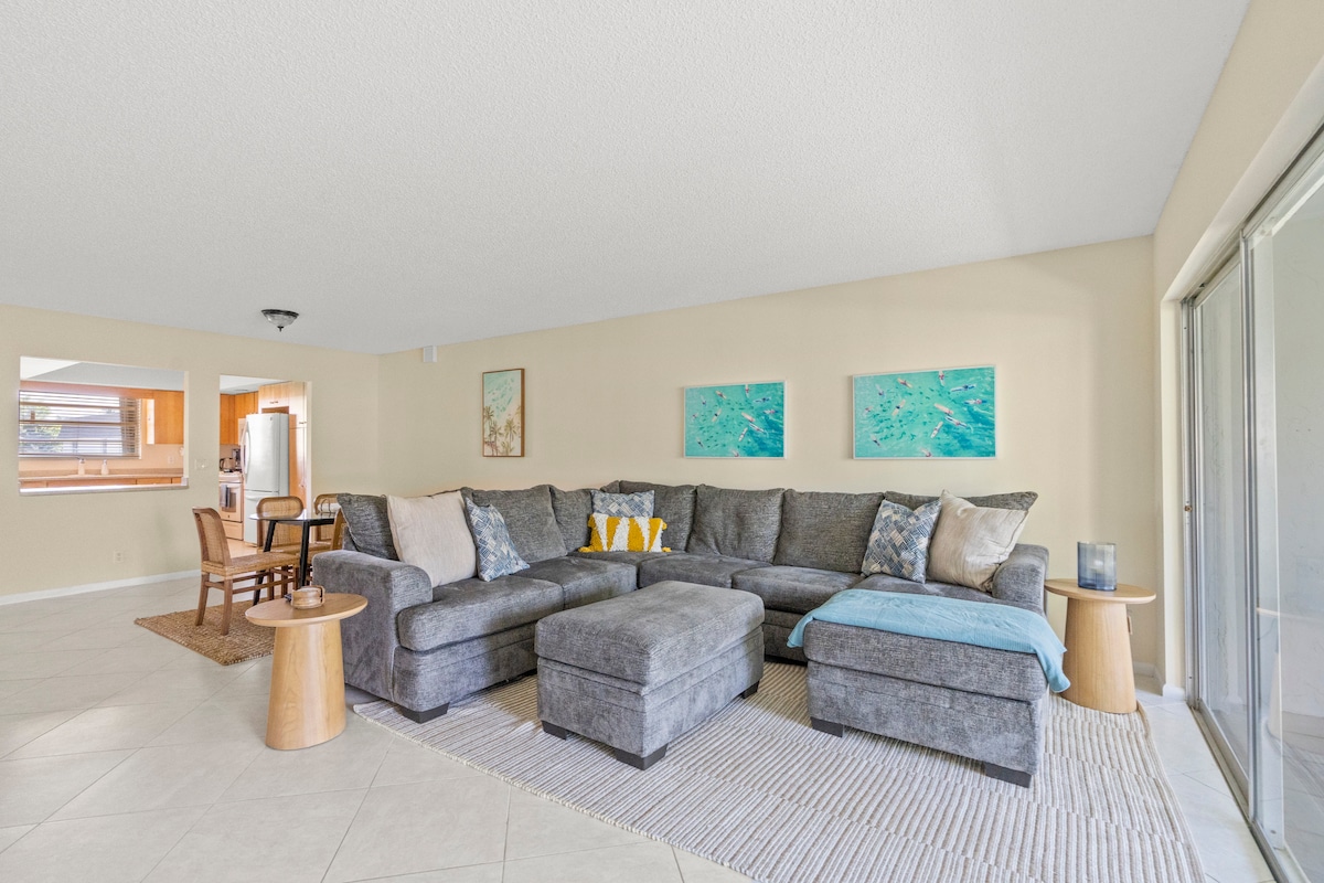 A spacious living area is furnished with a large gray sectional sofa adorned with decorative pillows. Complementing the seating are light wooden side tables. Ocean-themed artwork hangs on the walls, and natural light fills the room through large sliding glass doors leading to the patio.