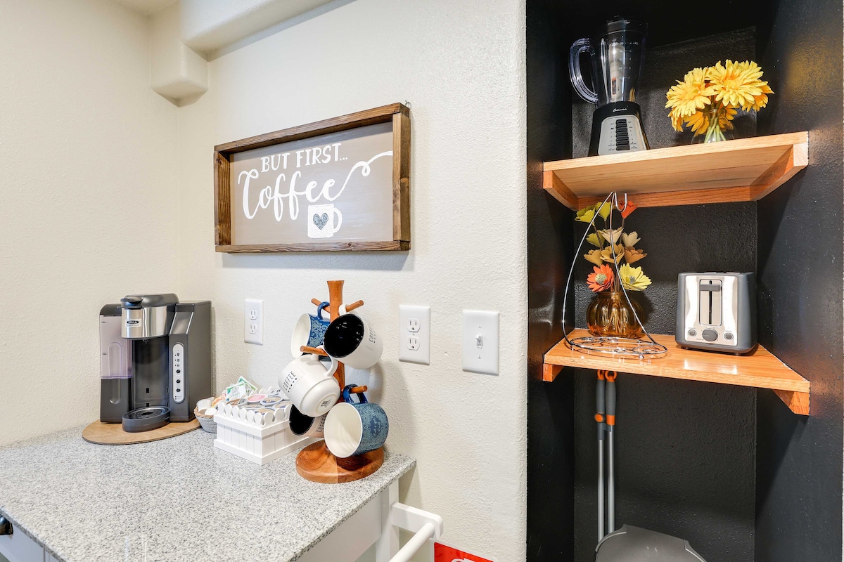A well-equipped kitchenette displays a coffee maker beside a small dish rack filled with colorful mugs. A blended countertop offers a modern finish. A shelf features a blender, toaster, and a vase of sunflowers, enhancing the overall functionality of the space.