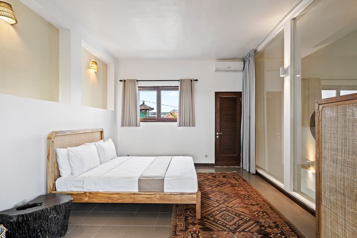 Chumba cha kulala kina mchanganyiko wa vitu vya kisasa na vya asili, vilivyo na kitanda kilicho na ubao wa kichwa wa rattan uliosukwa, mashuka meupe, mapambo madogo na mazingira angavu.