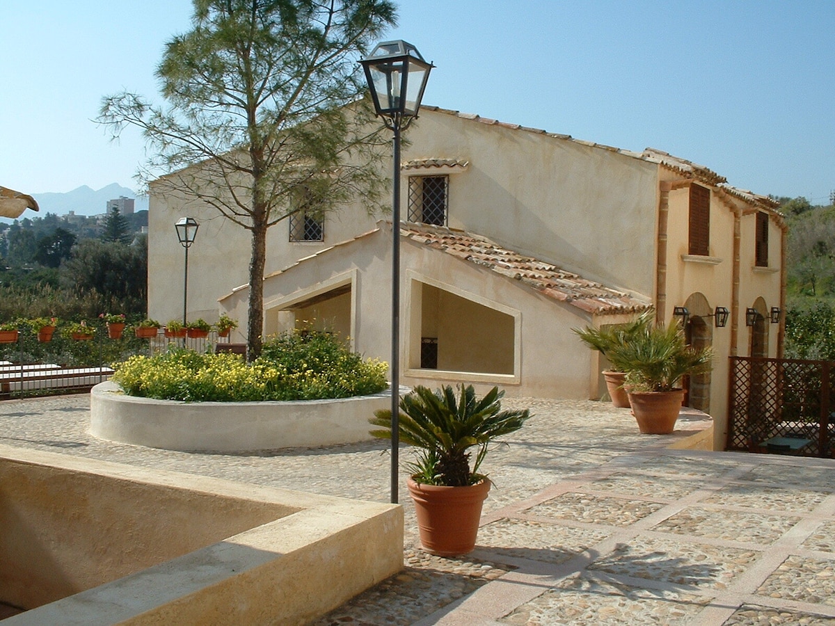 A charming farmhouse with a rustic stone facade is surrounded by greenery. The entrance features a circular flower bed with vibrant yellow blooms. Potted plants add a touch of nature to the area. Stone pathways and a lamp post enhance the welcoming outdoor space.