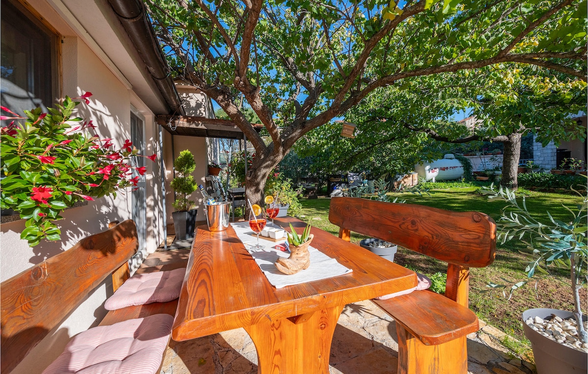 An outdoor dining area features a wooden table surrounded by bench seating. A table runner is arranged with a small centerpiece. Lush greenery and blooming flowers are visible, enhancing the tranquil atmosphere in the inviting space under the shade of a tree.