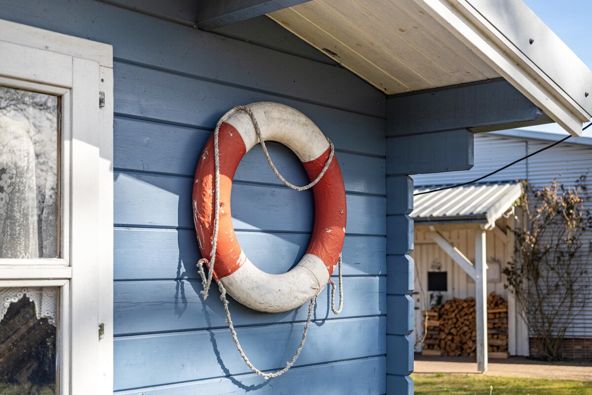 A life preserver is mounted on a blue wooden wall, featuring a white center and orange exterior. The surrounding area includes a visible wooden structure and neatly stacked firewood in the background, indicating a cozy and functional outdoor space.