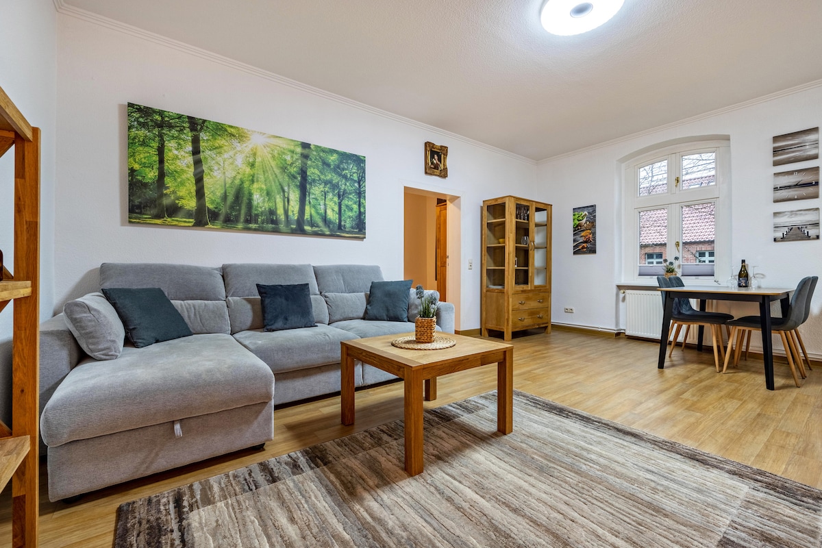 A welcoming living area features a spacious grey sofa adorned with cushions, complemented by a wooden coffee table. Natural light filters through the large windows, highlighting a dining table and wooden shelving. Artwork depicting a forest scene adds a touch of nature to the calm atmosphere.