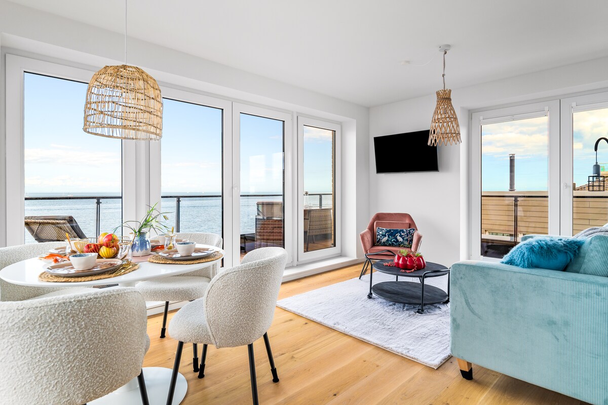 A light-filled living area features large glass doors providing views of the waterfront. A round dining table is set for four, while a cozy armchair and a comfortable sofa offer seating. A flat-screen TV is mounted on the wall, and soft neutral tones are used throughout.