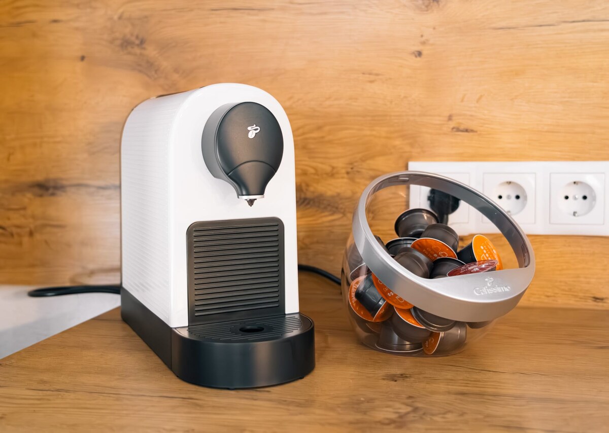 A sleek coffee machine is positioned on a wooden countertop, accompanied by a transparent storage container filled with coffee capsules. The machine features a simple, modern design with a single dispensing spout.