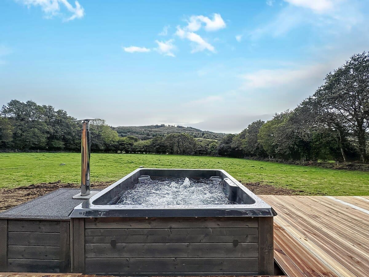 An outdoor jacuzzi is set on a wooden deck, surrounded by lush greenery and open fields. The water appears inviting as it bubbles and splashes, with rolling hills visible in the distance under a clear sky.