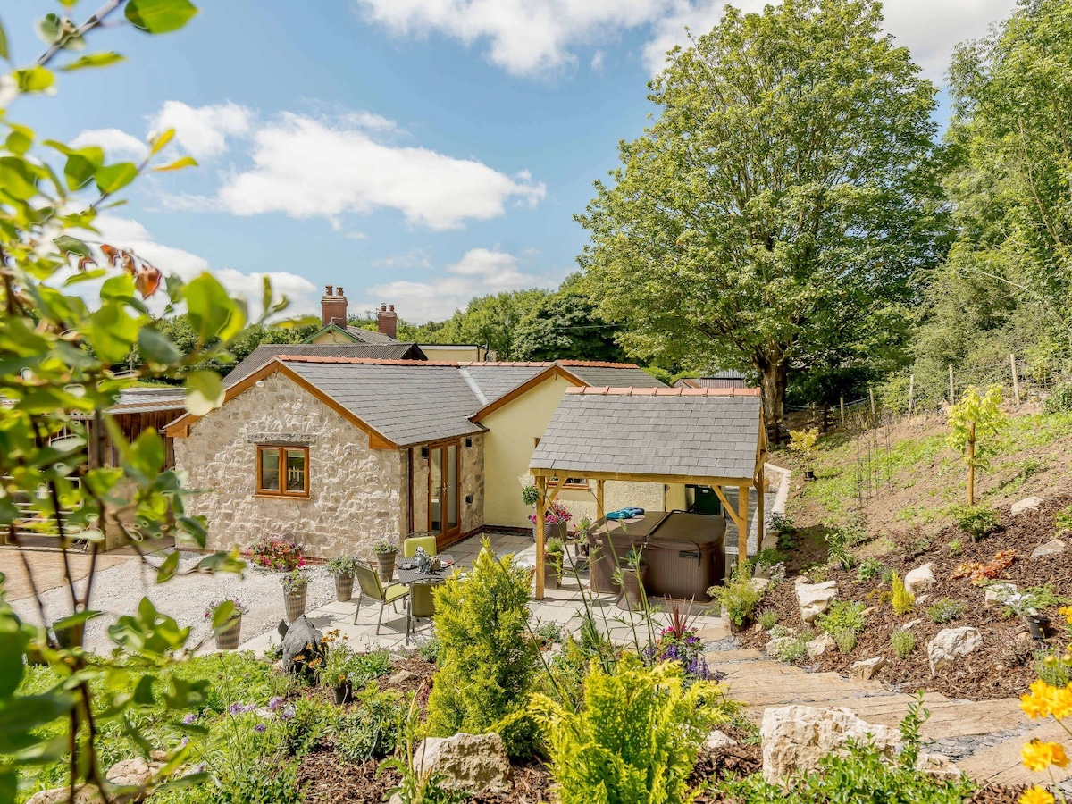 A charming stone cottage is nestled amidst lush greenery, featuring a patio area with seating and a hot tub. Vibrant flowers and shrubs surround the property, while a spacious tree provides natural shade. The cottage's exterior exhibits a warm colour palette with large windows for natural light.