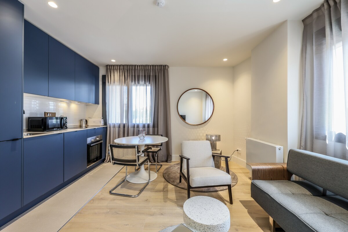 A modern living-dining area is shown, featuring an open kitchen along one wall with dark cabinetry. A circular dining table is set for four, and a round mirror reflects light. Seating includes a comfortable armchair and a gray sofa, complemented by soft curtains covering the large window.