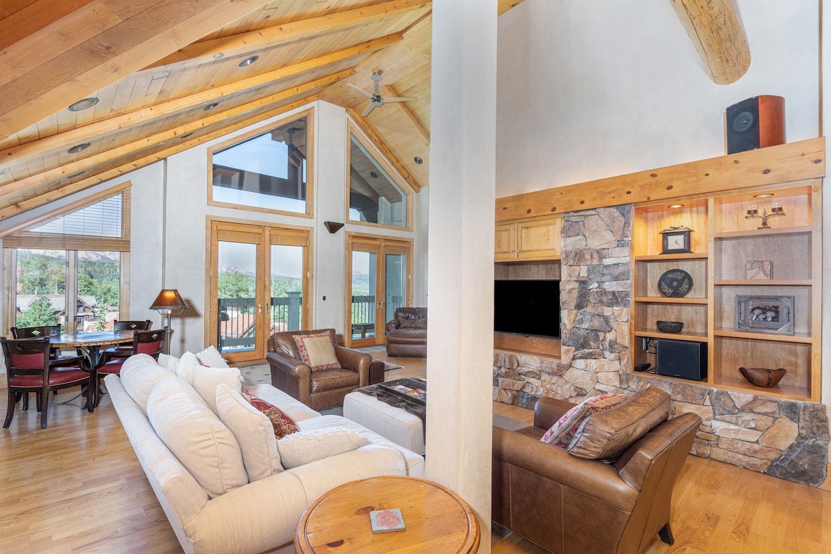 A spacious living area features a central stone fireplace and vaulted wood ceilings. Large windows provide abundant natural light and views of the outdoors. The seating arrangement includes a plush, white sofa and leather armchairs, with a wooden table set for dining in the background.