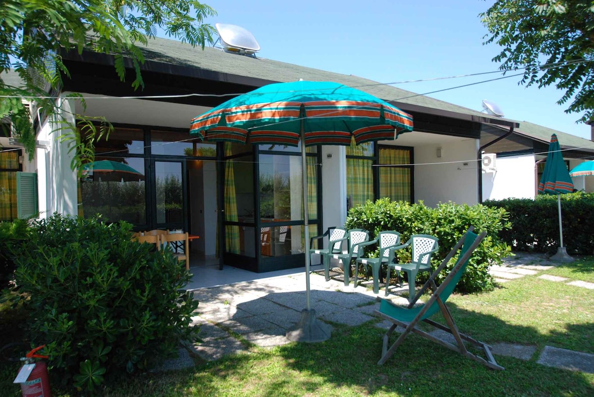 A functional outdoor area features a large umbrella providing shade over a table and chairs, enhancing the space for relaxation. Lush greenery surrounds the area, while additional seating options are visible, offering a welcoming environment for outdoor enjoyment.