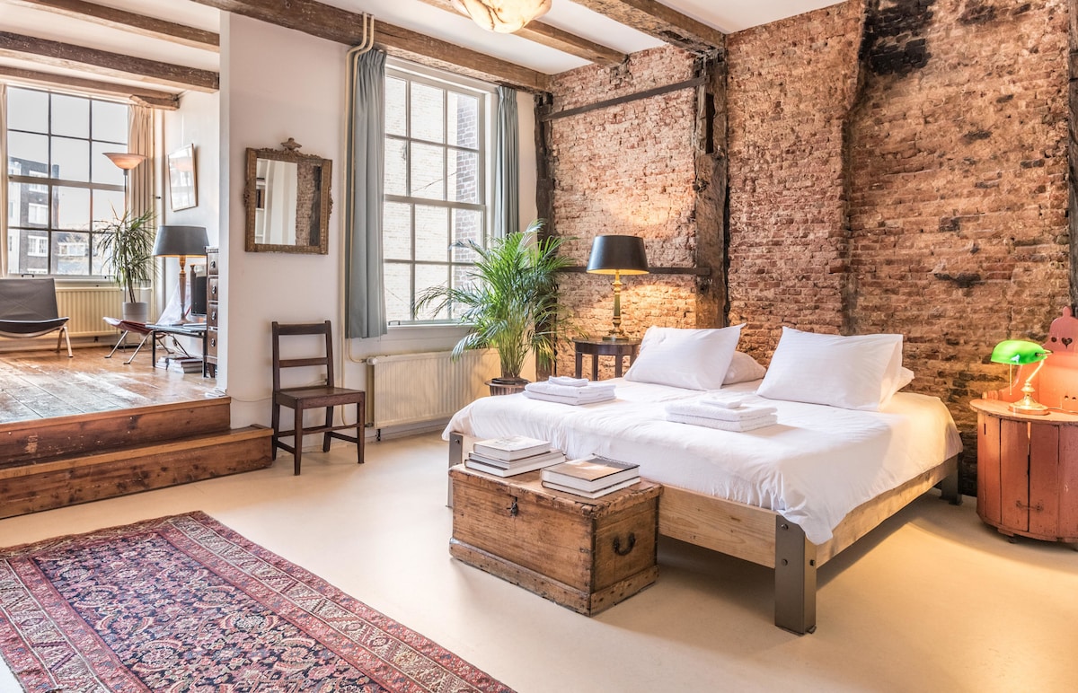 The spacious room features exposed oak beams and an authentic brick wall. A double bed with two separate mattresses is centered, accompanied by a vintage trunk at its foot. A seating area with a chair and a lamp is present, with natural light streaming through large windows.