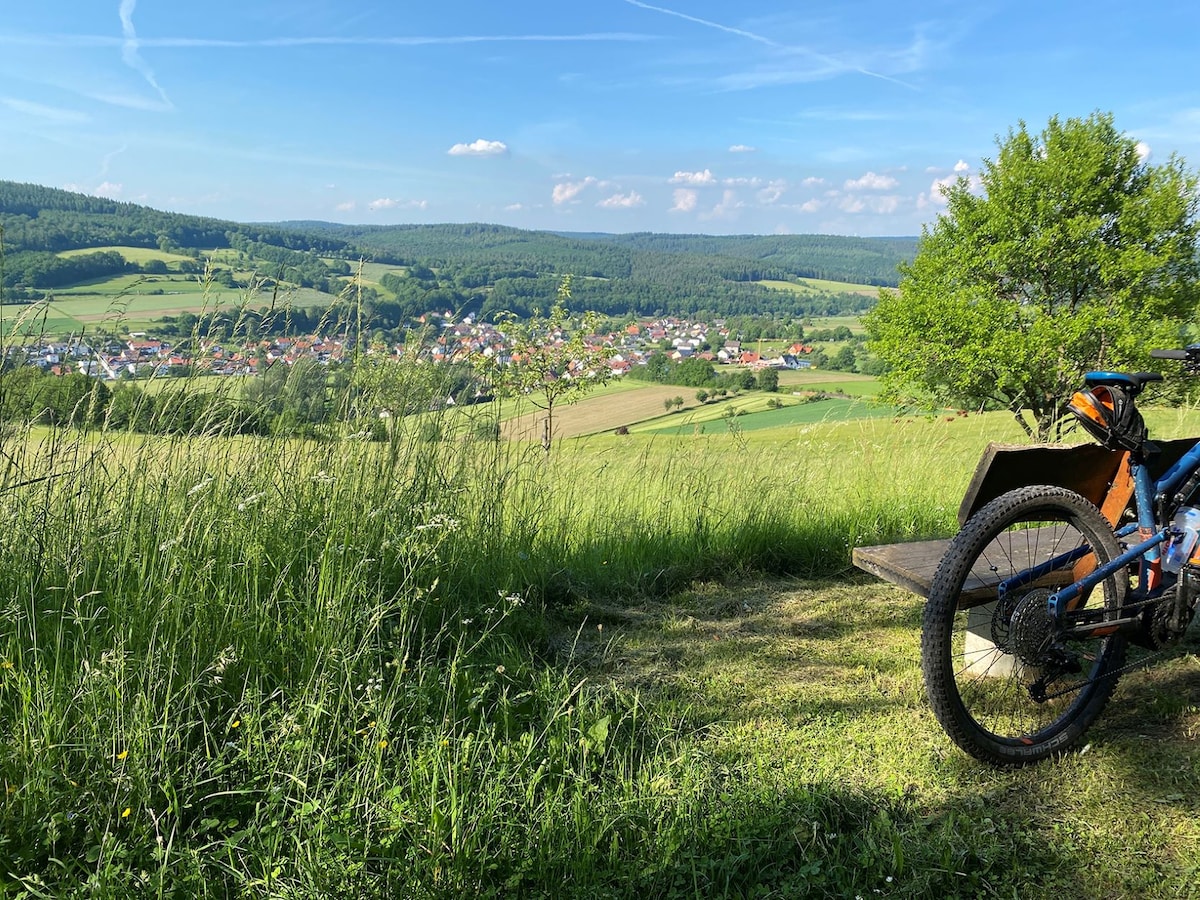 A scenic view is captured from a grassy hilltop, overlooking a charming village nestled in a lush green valley. A bicycle rests in the foreground, surrounded by tall grasses, while the expansive sky features scattered clouds and sunlight.