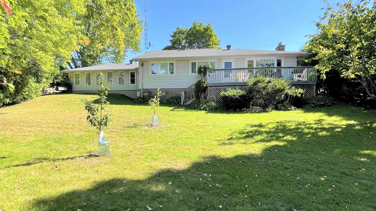 The exterior of a cozy cottage showcases a spacious lawn surrounded by trees, providing a sense of privacy. A large deck extends from the cottage, and a sunroom is visible to the left, all under a clear blue sky.