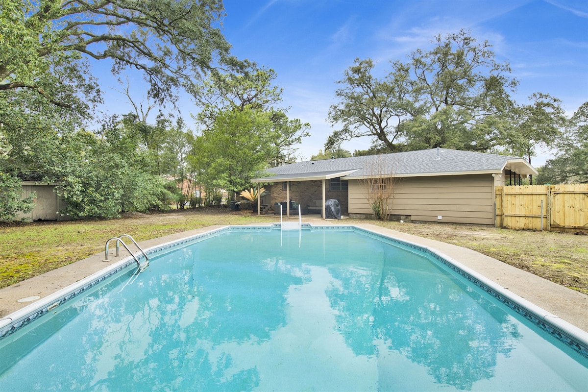A refreshing pool is situated in a spacious yard, surrounded by greenery. Comfortable lounge chairs are positioned nearby, offering a place to relax. The pool features steps leading in, and a tranquil atmosphere is presented by the well-maintained landscaping and the covered backyard area.