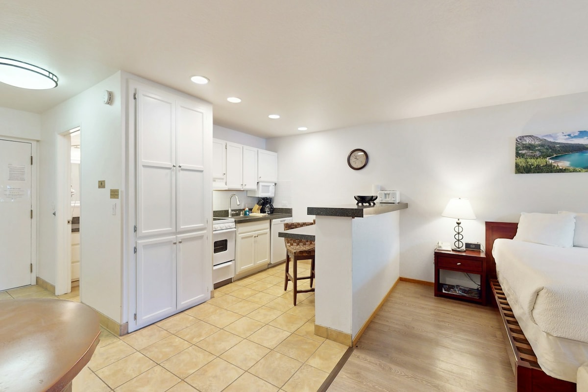 The studio's open layout features a well-equipped kitchen with white cabinetry and modern appliances. A kitchen bar with seating is positioned beside a small dining table. Natural light fills the space, highlighting the cozy sleeping area and tasteful mountain-themed decor.