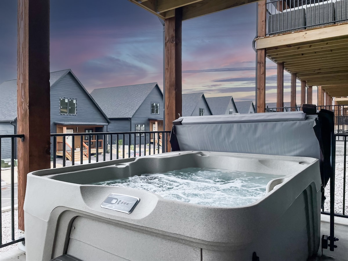 A private hot tub is situated on a covered deck, surrounded by a railing. Bubbles can be seen gently rising in the water. In the background, multiple vacation homes are visible beneath a colorful, twilight sky.