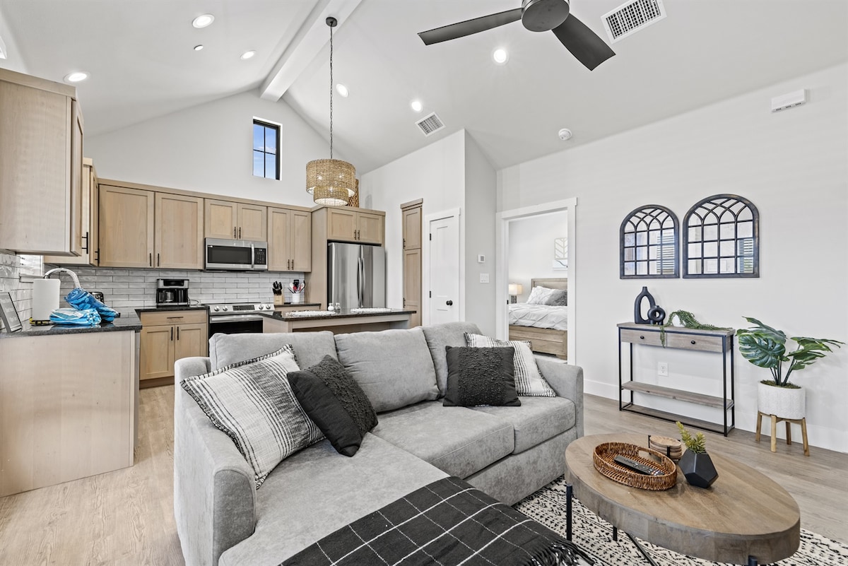 An open living area features a spacious gray sectional sofa, accompanied by a round wooden coffee table and a decorative plant. The kitchen, designed with light wood cabinets and stainless steel appliances, is visible in the background, enhancing the airy feel of the space.