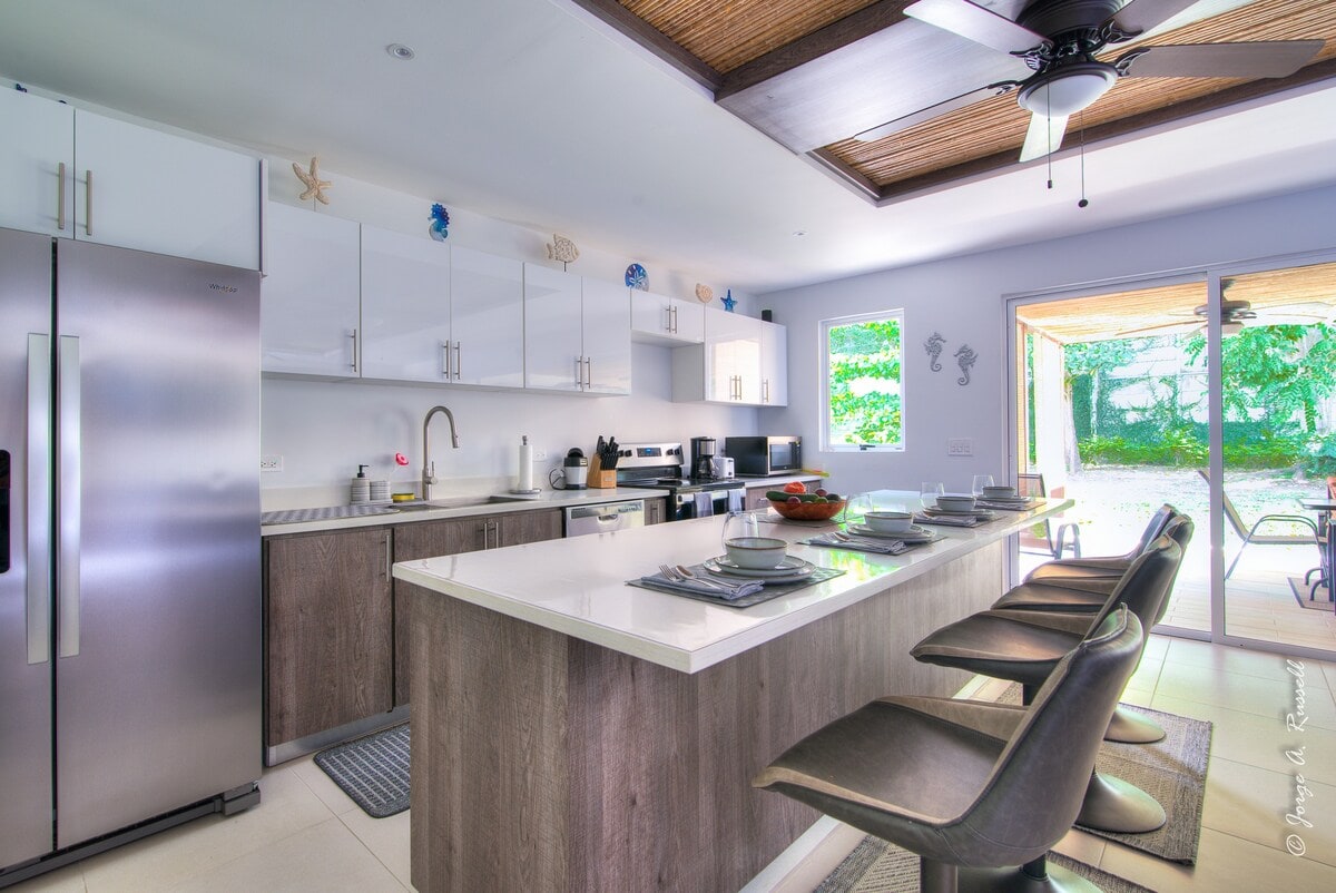 A modern kitchen features sleek cabinetry and stainless-steel appliances, including a refrigerator and a coffee maker. A spacious island with stools provides seating, while large windows allow natural light to fill the area. A door opens to an outdoor space, enhancing the inviting setting.