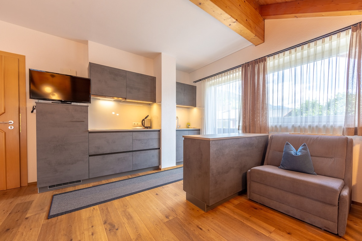 The open-plan living area features a modern kitchen with sleek cabinetry and integrated appliances. A comfortable sofa is positioned nearby, complemented by a decorative pillow. Natural light streams through large windows covered with sheer curtains, illuminating the warm wooden flooring.