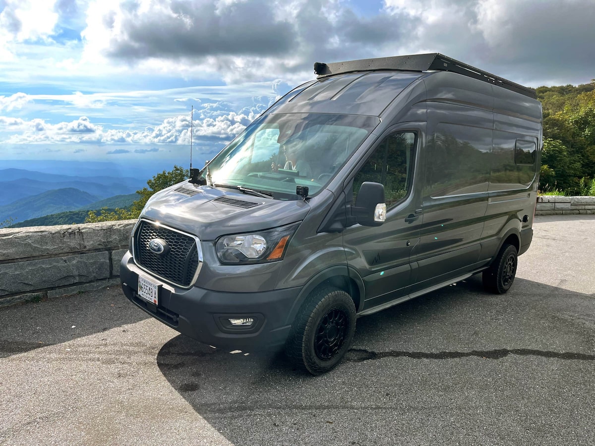 The camper van is parked on a concrete surface, featuring a sleek grey exterior. Off-road tires are visible, enhancing its rugged appeal. In the background, expansive mountain vistas and a cloudy sky can be seen, showcasing the vehicle's suitability for outdoor adventures.
