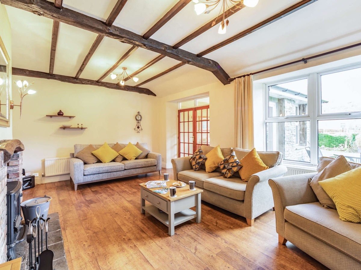 A spacious lounge features exposed wooden beams and warm lighting. Comfortable seating is arranged around a central coffee table, and decorative cushions add a touch of colour. A wood burner is positioned against the wall, with a set of French doors leading to the outdoor area.