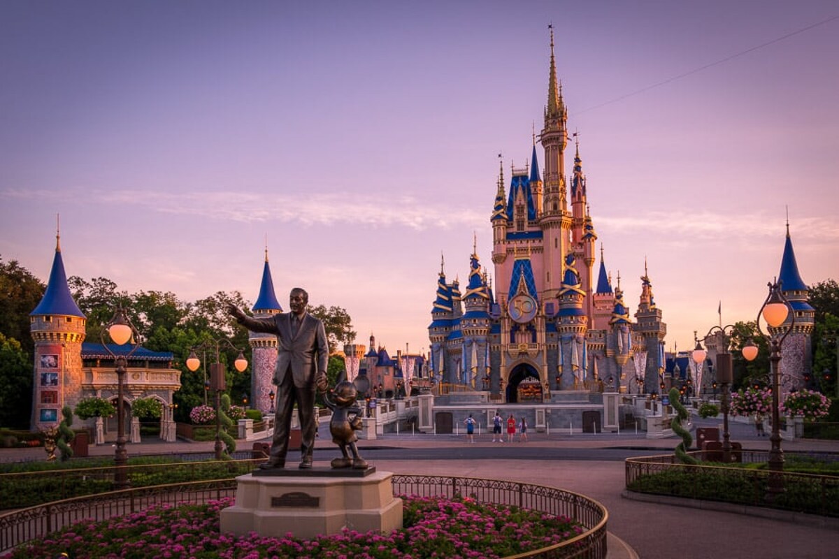 A majestic castle stands at the center, adorned with vibrant blue and gold spires. Surrounding the castle, lush greenery and colorful flower arrangements can be seen, while vintage lampposts provide soft illumination. A statue of a man and a mouse is prominently featured in the foreground.