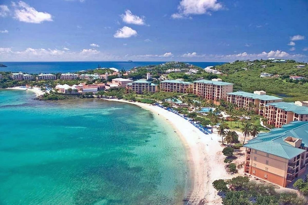 An aerial view captures the expansive beachfront property with soft sandy shores and turquoise waters. The resort buildings, featuring pale earth tones and teal roofs, are set against lush green hills, creating a serene coastal atmosphere under a clear blue sky.