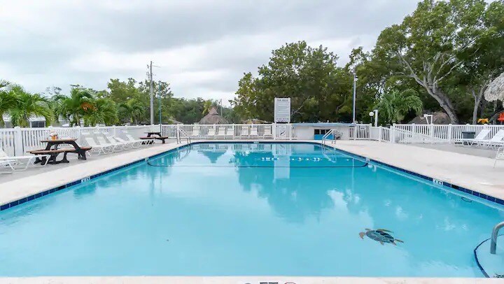 A heated outdoor swimming pool is surrounded by lounge chairs and picnic tables. Lush greenery and palm trees provide shade, while a thatched roof structure is visible in the background. The pool's clear blue water reflects the overcast sky.