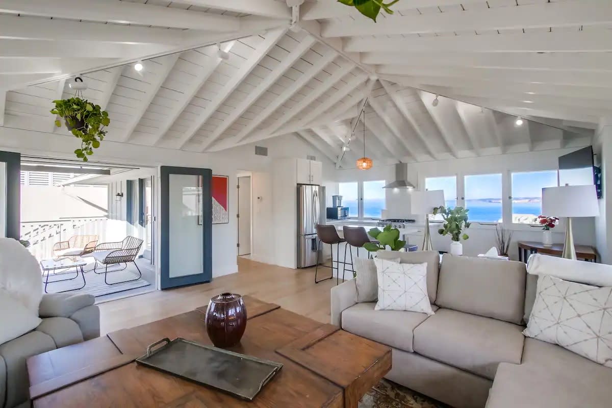 The open living area showcases high white beamed ceilings, with plush seating arranged around a wooden coffee table. A modern kitchen is visible, featuring stainless steel appliances. Bright views of the ocean are captured through multiple windows that illuminate the space.