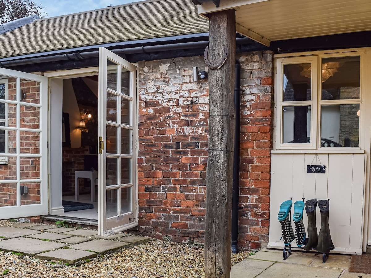 An entrance area features double doors opening to a warm indoor space. Exposed brick walls and wooden beams enhance the rustic feel. On the side, a pair of wellies is neatly arranged, indicating a connection to nature and outdoor activities.