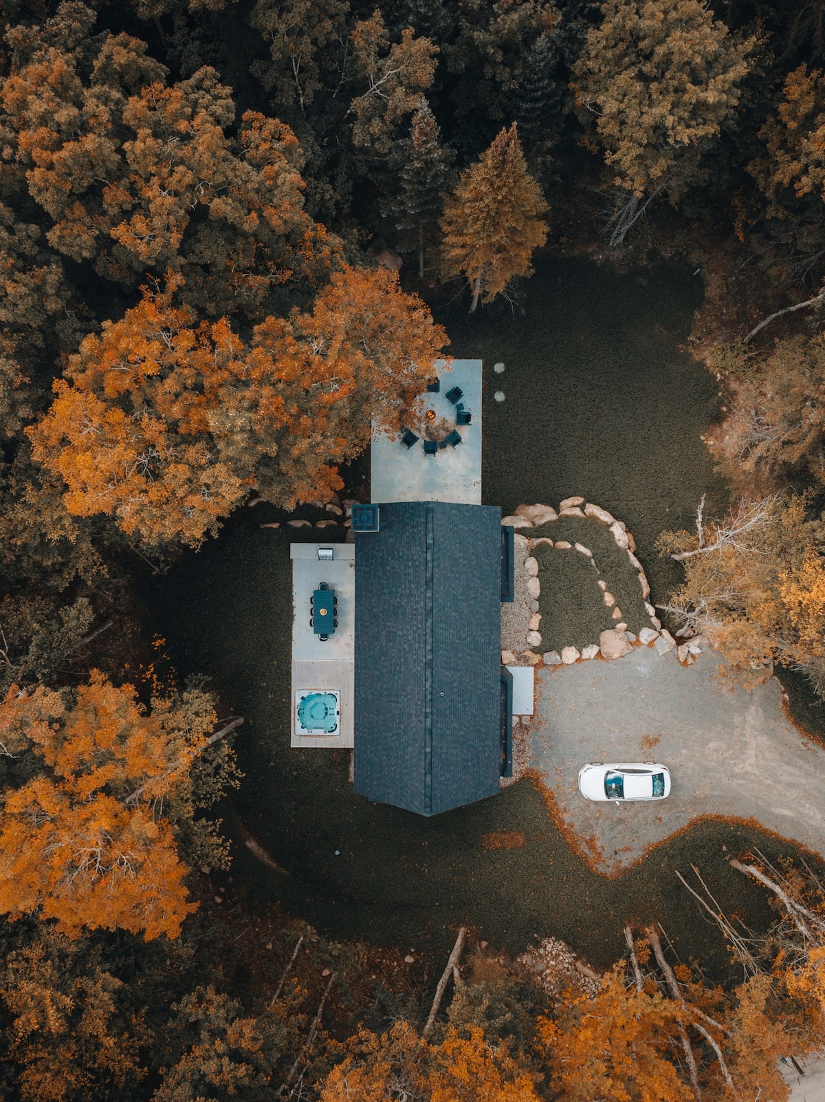 An aerial view of a modern cabin surrounded by vibrant autumn foliage. A hot tub and outdoor dining area are positioned next to the cabin. A gravel pathway leads to the outdoor space, with a parking area visible on one side.