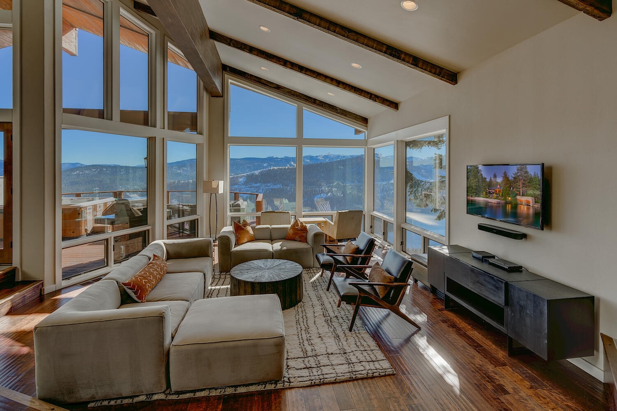 A spacious living area is illuminated by natural light from large windows, offering panoramic views of the mountains. A comfortable sectional sofa surrounds a modern coffee table, while two accent chairs complement the room. A flat-screen television is mounted on the wall, adding functionality to the space.