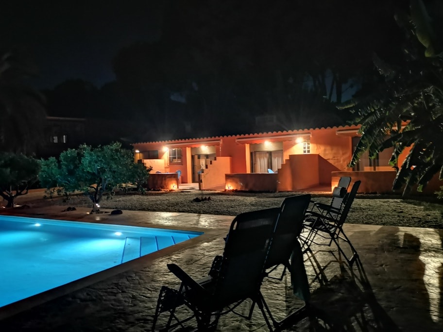 The outdoor area features a well-lit poolside scene, with a pool reflecting soft blue lights. Several black lounge chairs are positioned near the pool, and the warm glow emanates from the nearby building, surrounded by lush greenery and palm trees.