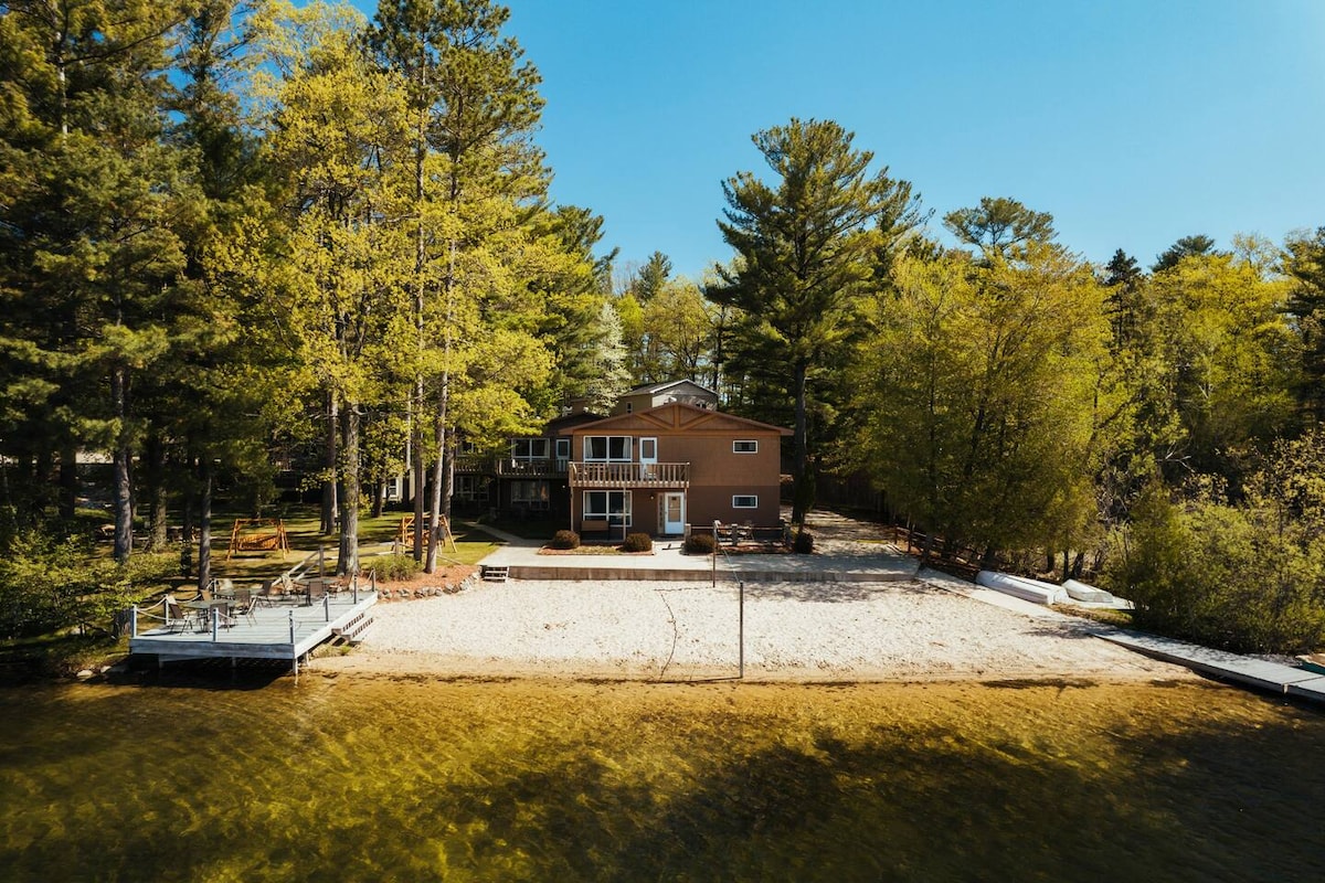 A view of the lakefront resort highlights the sandy beach area, surrounded by tall trees. The two-story building features multiple balconies and large windows, enhancing the inviting lakeside atmosphere. A dock extends into the water, providing easy access to the lake.