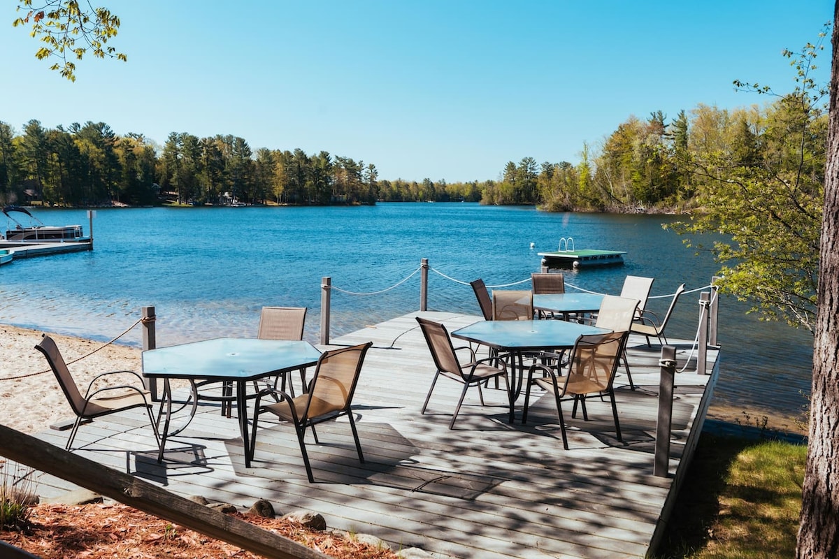 A spacious dock extends over the water, featuring several round glass-top tables surrounded by comfortable seating. Sunlight reflects off the calm lake, framed by a backdrop of trees and a sandy beach. A swimming platform is visible in the distance.
